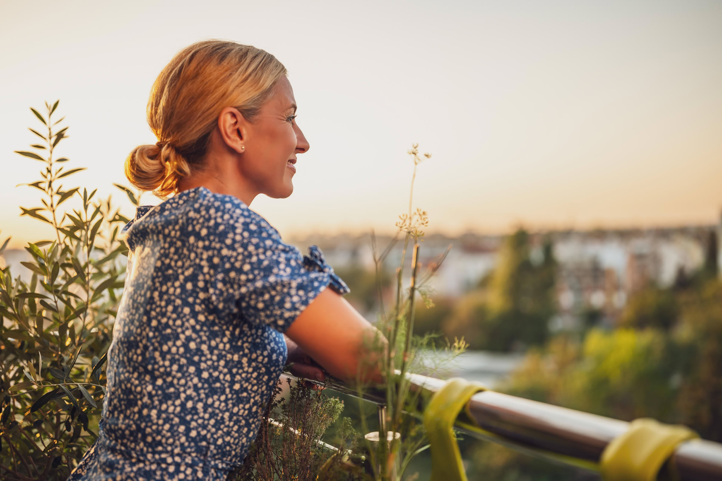 Magnolia Woman with blonde hair in a blue dress stands on a balcony, smiling and looking at a sunset over a green landscape.
