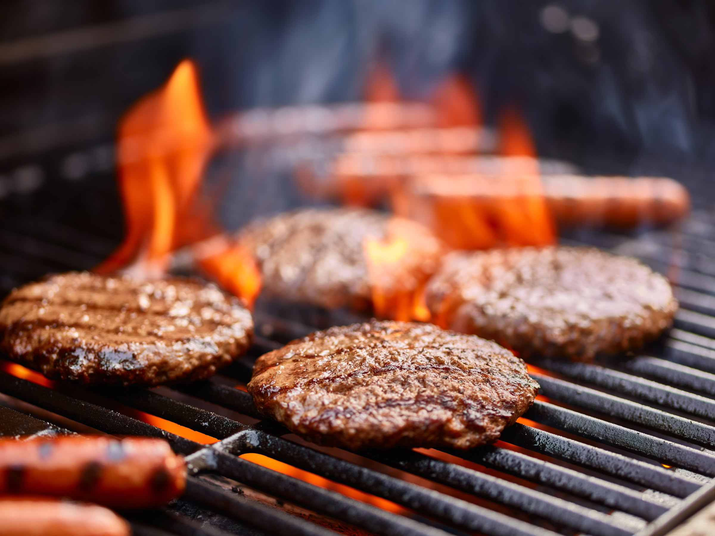 Magnolia Four burger patties and hot dogs are grilling over open flames on a barbecue grill.