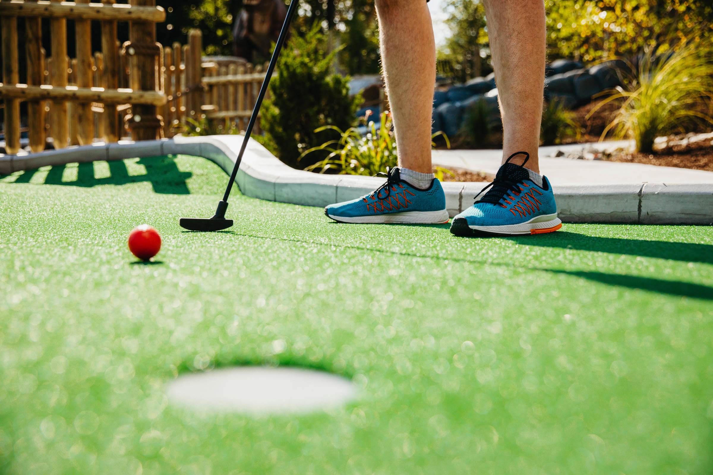 Magnolia Person in blue sneakers playing mini-golf, holding a putter near a red ball on a green course, aiming for the hole.