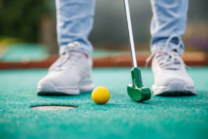 Magnolia Person wearing white sneakers about to putt a yellow golf ball toward a hole on a green mini-golf course.