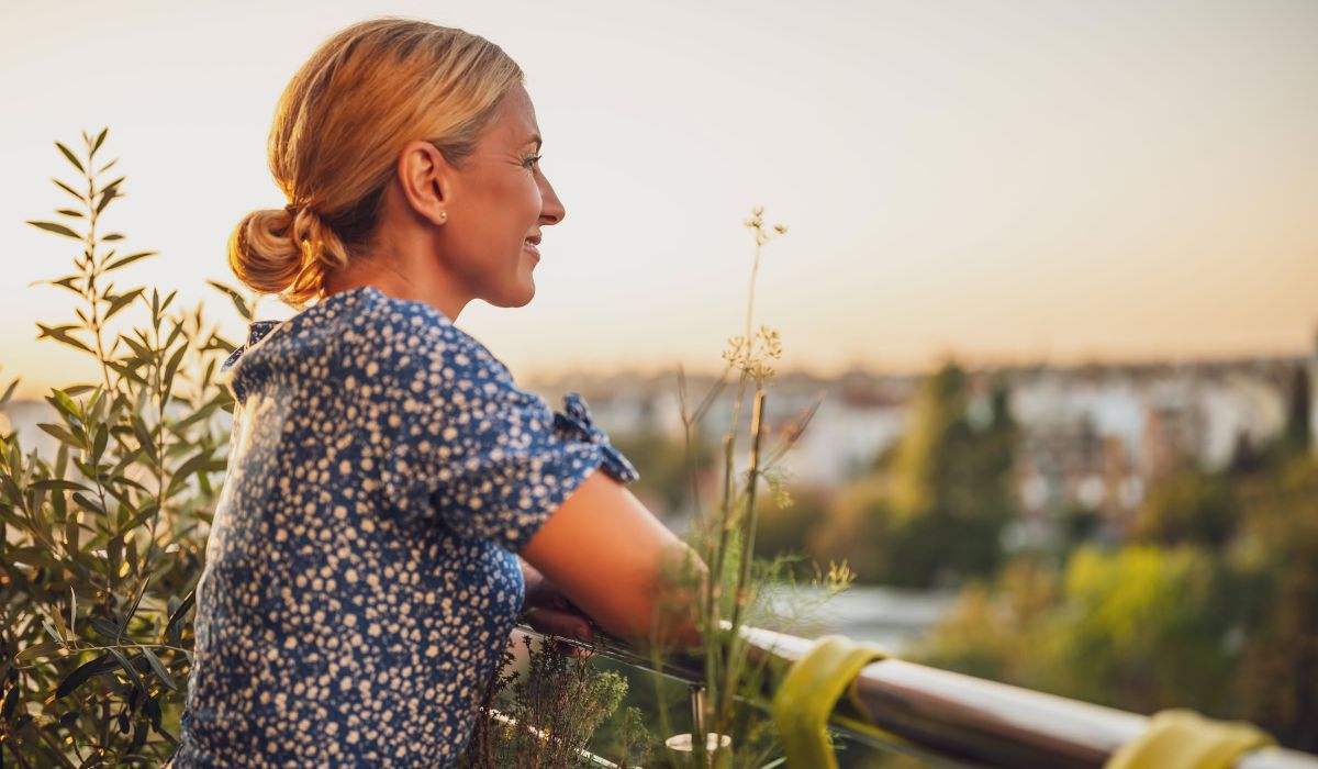 Magnolia Woman with blonde hair in a blue dress stands on a balcony, smiling and looking at a sunset over a green landscape.