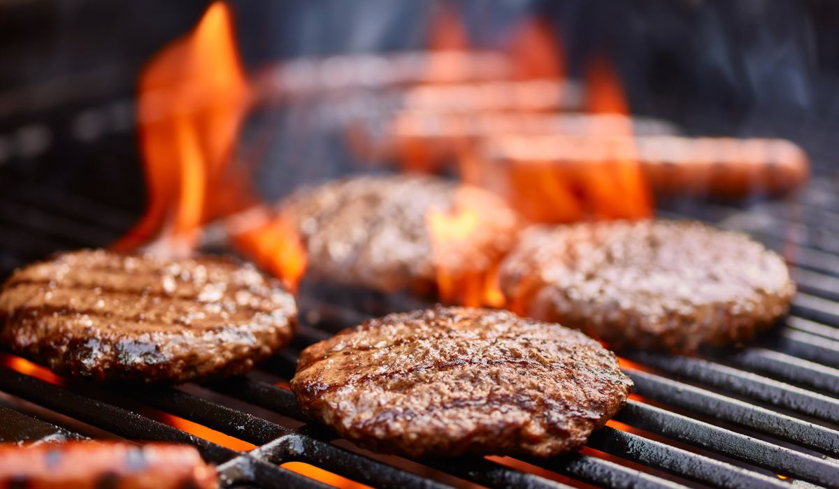 Magnolia Four burger patties and hot dogs are grilling over open flames on a barbecue grill.
