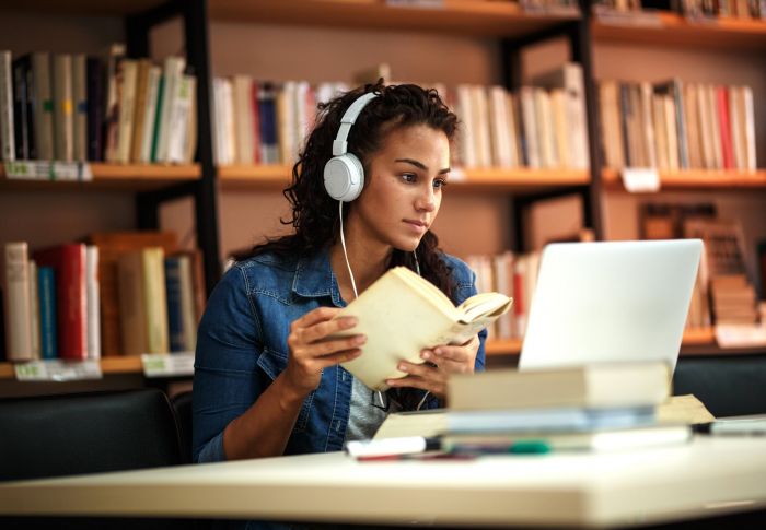 Magnolia Woman wearing headphones reads a book and looks at a laptop in a library.